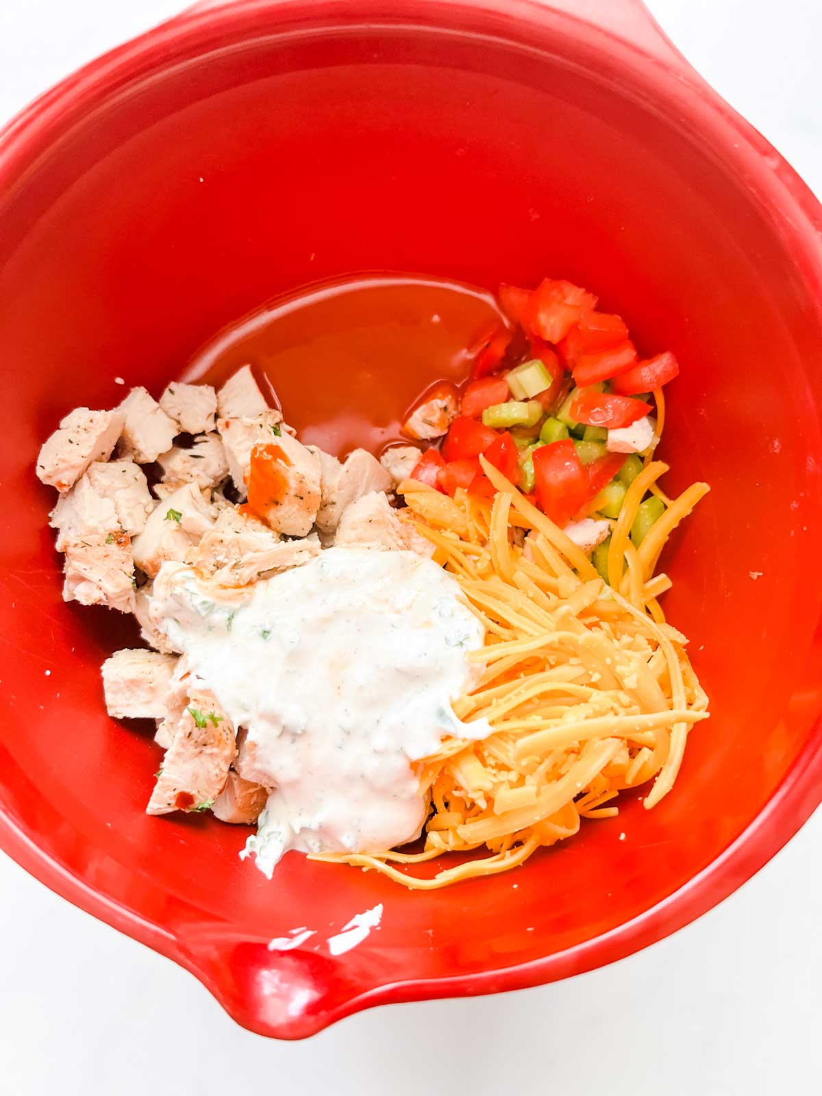 Photo of cubed chicken, celery, tomato, shredded cheddar, hot sauce, and ranch in a red bowl.