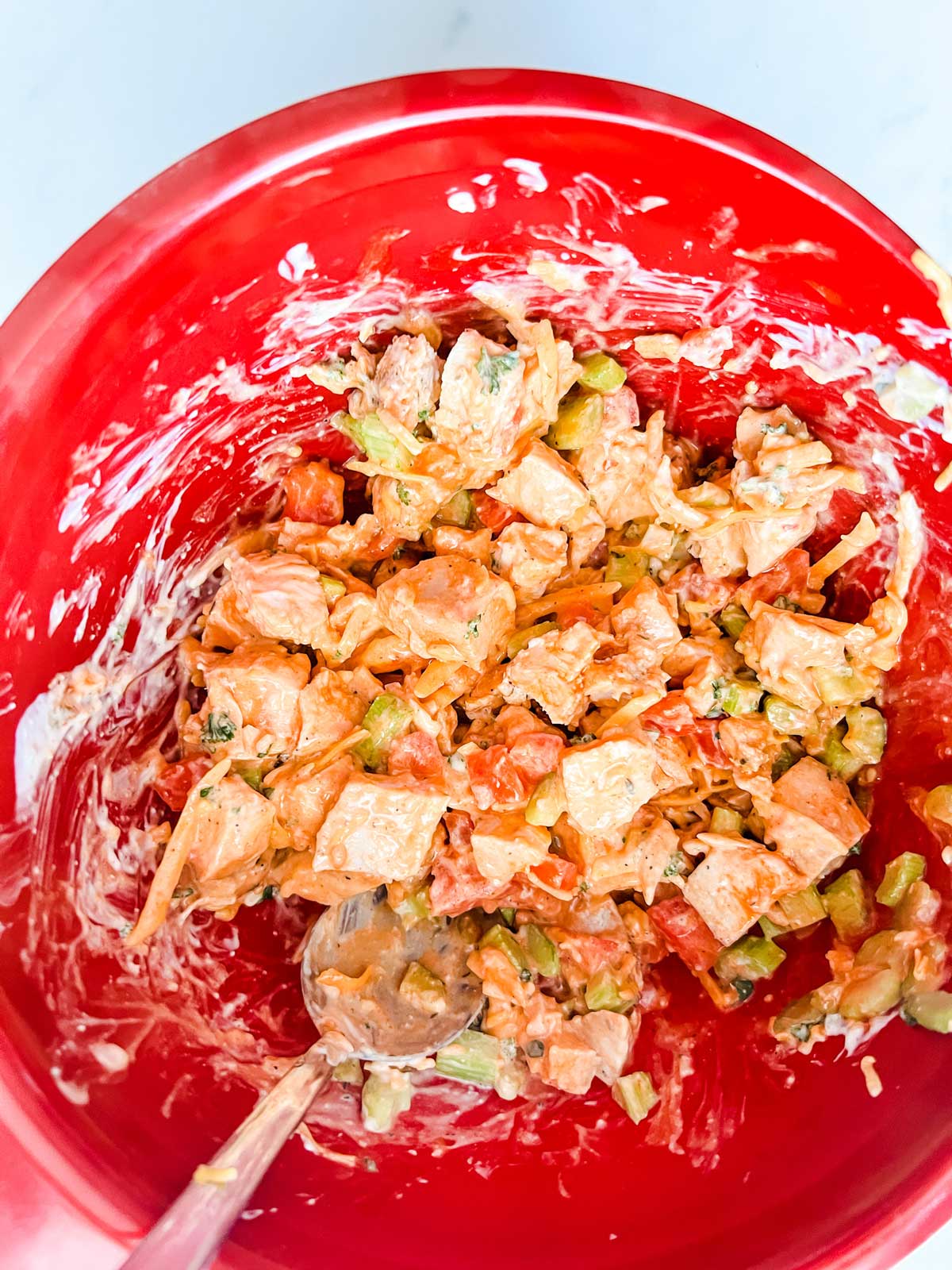 Photo of cubed chicken, celery, tomato, shredded cheddar, hot sauce, and ranch that have been mixed togetherin a red bowl.