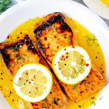 Close up photo of keto salmon in a plate of butter garnished with lemon, dill, and crushed red pepper flakes.