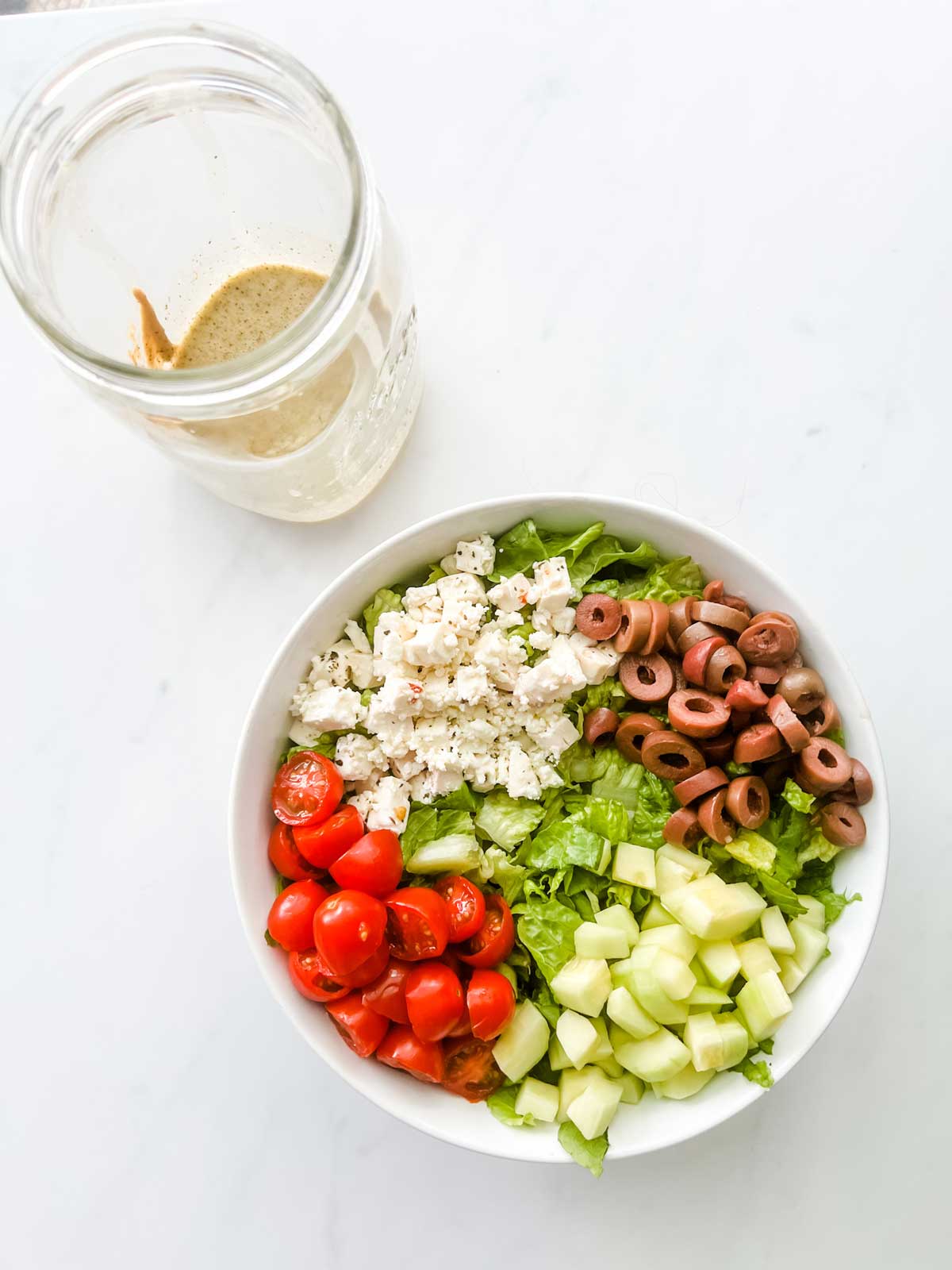 A bowl with lettuce, olive, cucumber, feta, and tomato with a mason jar of Greek salad dressing sitting next to it.