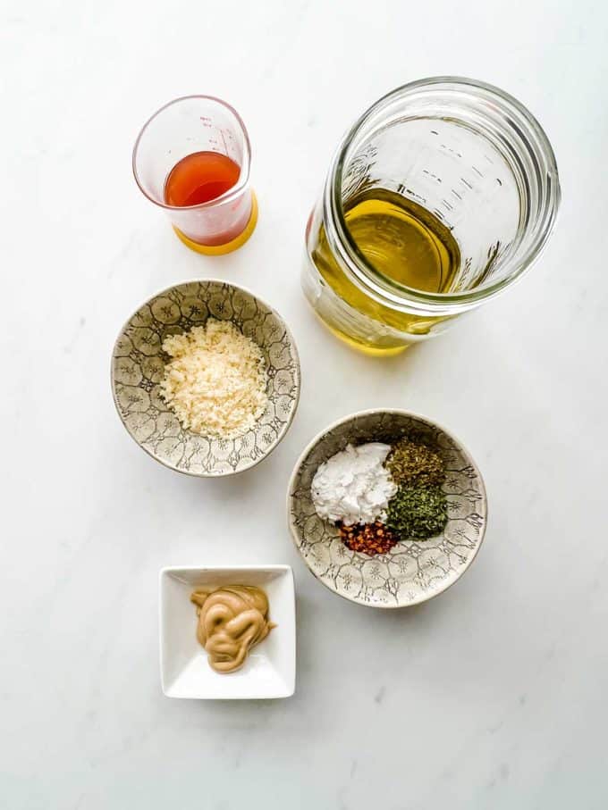 Overhead photo of a jar of oil, containers of vinegar, parmesan, sweetener, seasonings, and dijon.