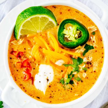 Close up square overhead photo of keto white chicken chili in a white crock garnished with lime, cheese, jalapeno, tomato, sour cream, and cilantro.