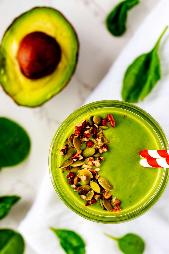 Overhead photo of a keto green smoothie garnished with pumpkin seeds and pecans and surrounded by spinach and avocado.