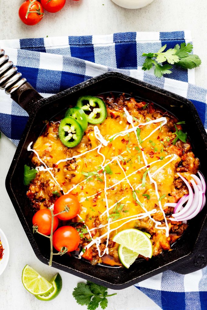 Overhead photo of a cast iron skillet with a cheesy taco skillet in it garnished with jalapeno, tomato, lime, and red onion.