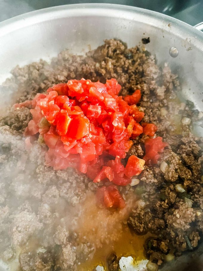A can of tomatoes added to a skillet of ground beef, vegetables, and seasonings.
