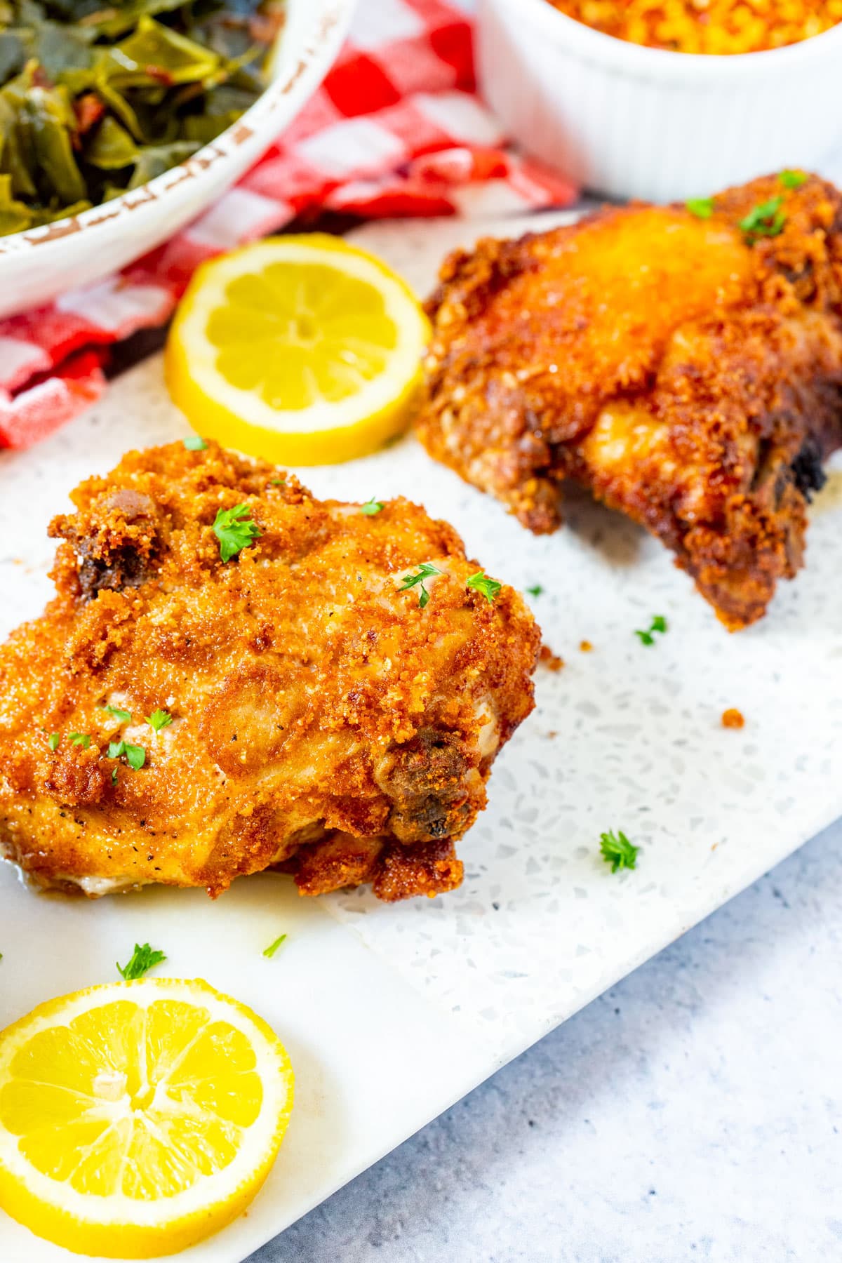 Two pieces of crispy fried chicken garnished with parsley are served on a white platter with lemon slices. Bowls of collard greens and corn are in the background on a red and white checkered cloth.