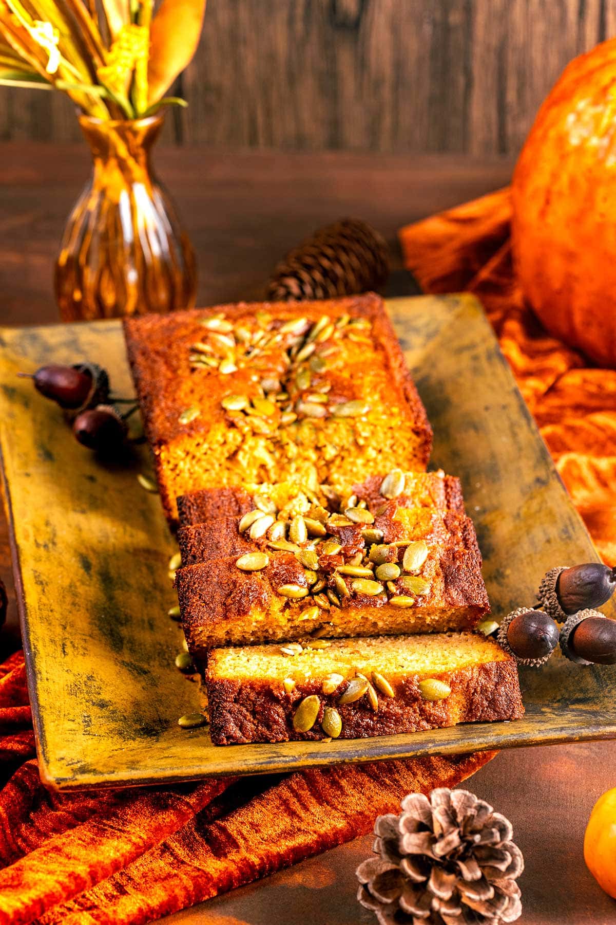 A loaf of pumpkin bread topped with seeds, sliced on a rustic rectangular plate. Autumn decorations like pinecones, acorns, and a pumpkin surround the bread on a wooden table with an orange cloth.