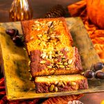 A golden-brown loaf of pumpkin bread with pumpkin seeds on top, partially sliced on a rustic yellow plate, surrounded by autumn-themed decor like pinecones and acorns.