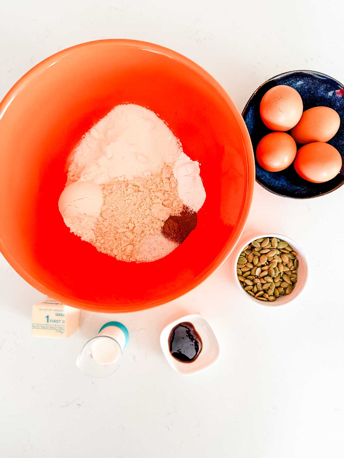A large orange bowl with almond flour and spices sits on a white surface, surrounded by four eggs in a blue bowl, a small bowl of pumpkin seeds, a stick of butter, a cup of milk, and a small bowl of dark liquid.