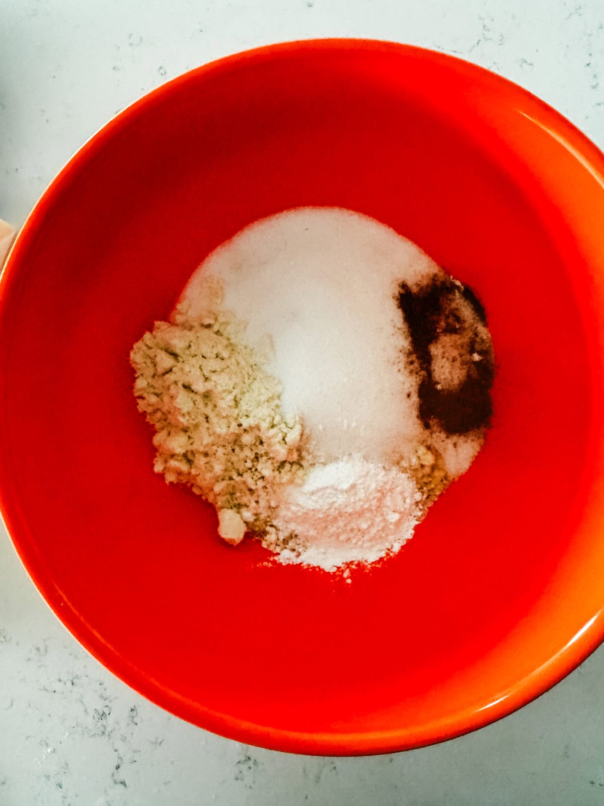 A bright orange bowl containing separate piles of dry ingredients, including sugar, flour, and spices, on a light-colored countertop.