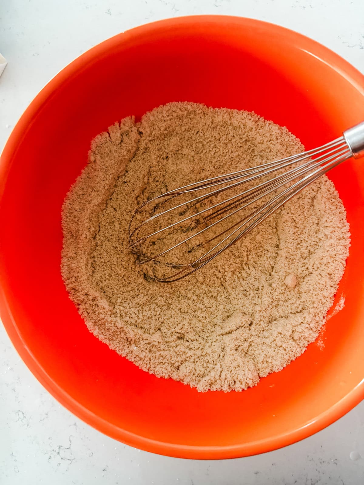 A metal whisk rests in a large orange bowl filled with a light brown, finely ground dry mixture on a white countertop.