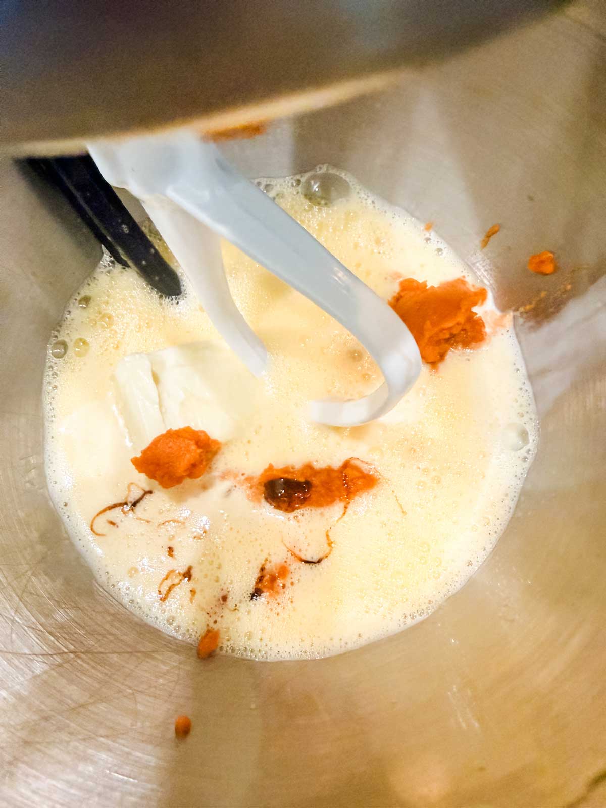 A stand mixer bowl with a paddle attachment, containing partially mixed ingredients including pumpkin puree, cream cheese, and frothy liquid.