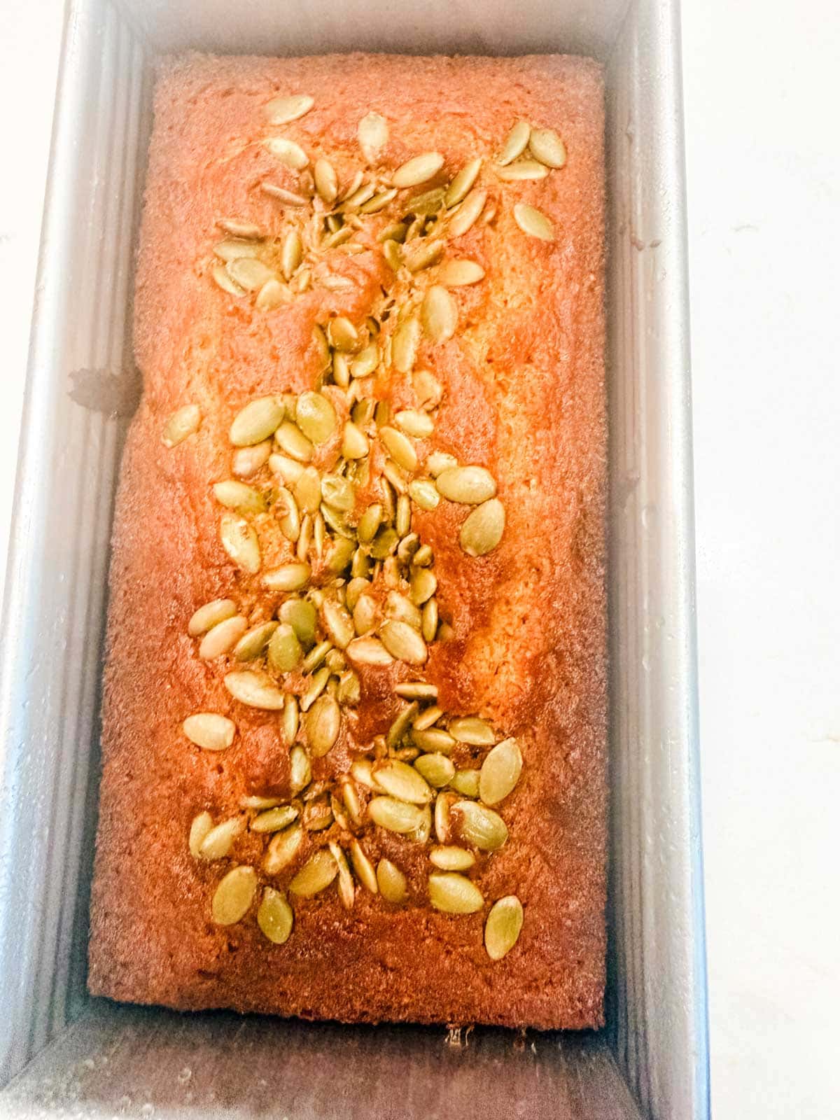 A loaf of golden-brown bread topped with scattered pumpkin seeds sits in a rectangular metal baking pan on a light-colored surface.