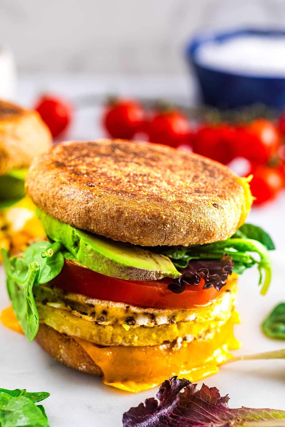 A breakfast sandwich on an English muffin with egg, melted cheddar cheese, tomato slice, leafy greens, and avocado, set on a white surface with cherry tomatoes in the background.