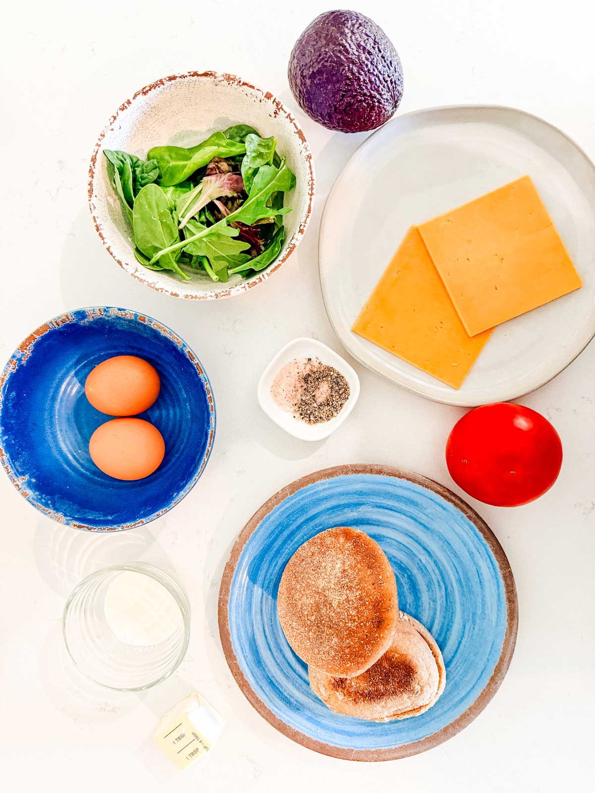 A flat lay of ingredients on a white surface: avocado, mixed greens, two cheddar cheese slices, two eggs, salt and pepper, a tomato, an English muffin sliced in half, cream, and a small piece of butter.
