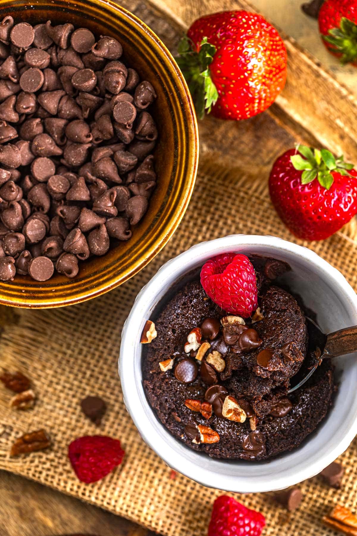 A bowl of chocolate chips sits next to a chocolate mug cake topped with pecans, chocolate chips, and a strawberry. Fresh strawberries are scattered on a rustic burlap surface.