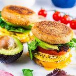 Two breakfast sandwiches with eggs, cheddar cheese, spinach, tomato, and avocado on whole wheat English muffins sit on a white surface, with half an avocado and some cherry tomatoes nearby.