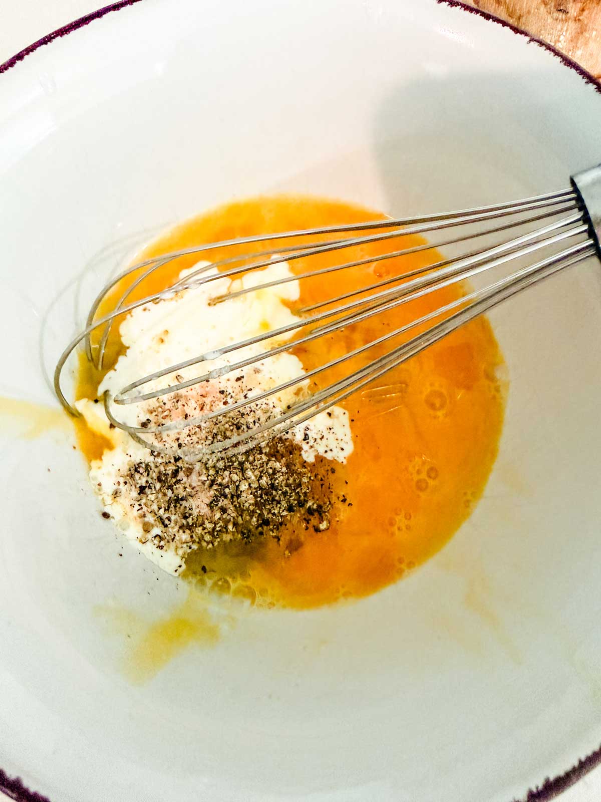 A metal whisk rests in a white bowl containing cracked eggs, cream, ground black pepper, and salt, ready to be mixed together.