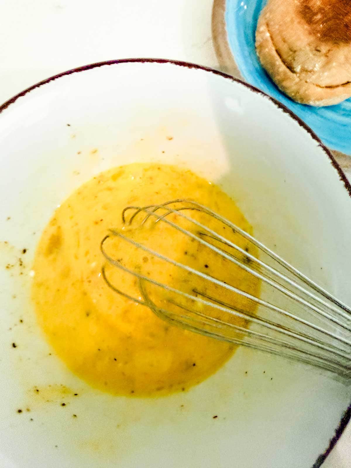 A metal whisk mixing beaten eggs with black pepper in a white bowl; part of a toasted bun on a blue plate is visible in the corner.