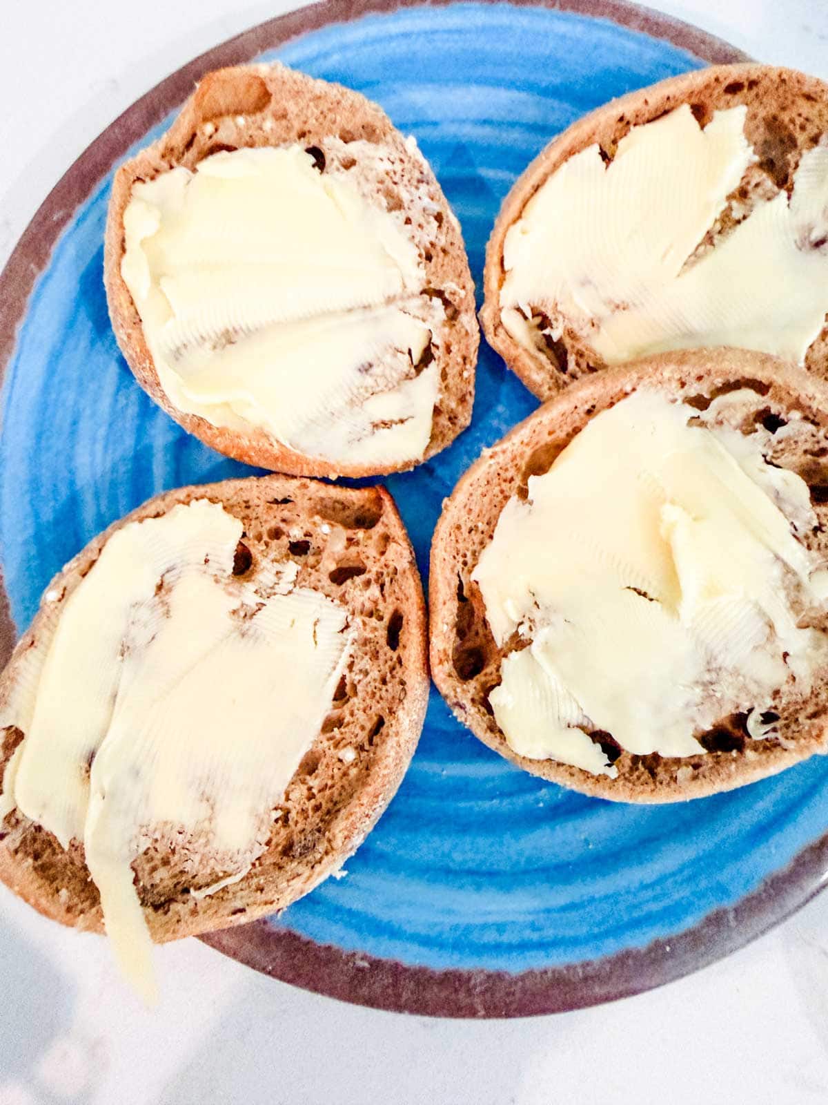 Four slices of brown english muffins topped with butter are arranged on a blue plate.