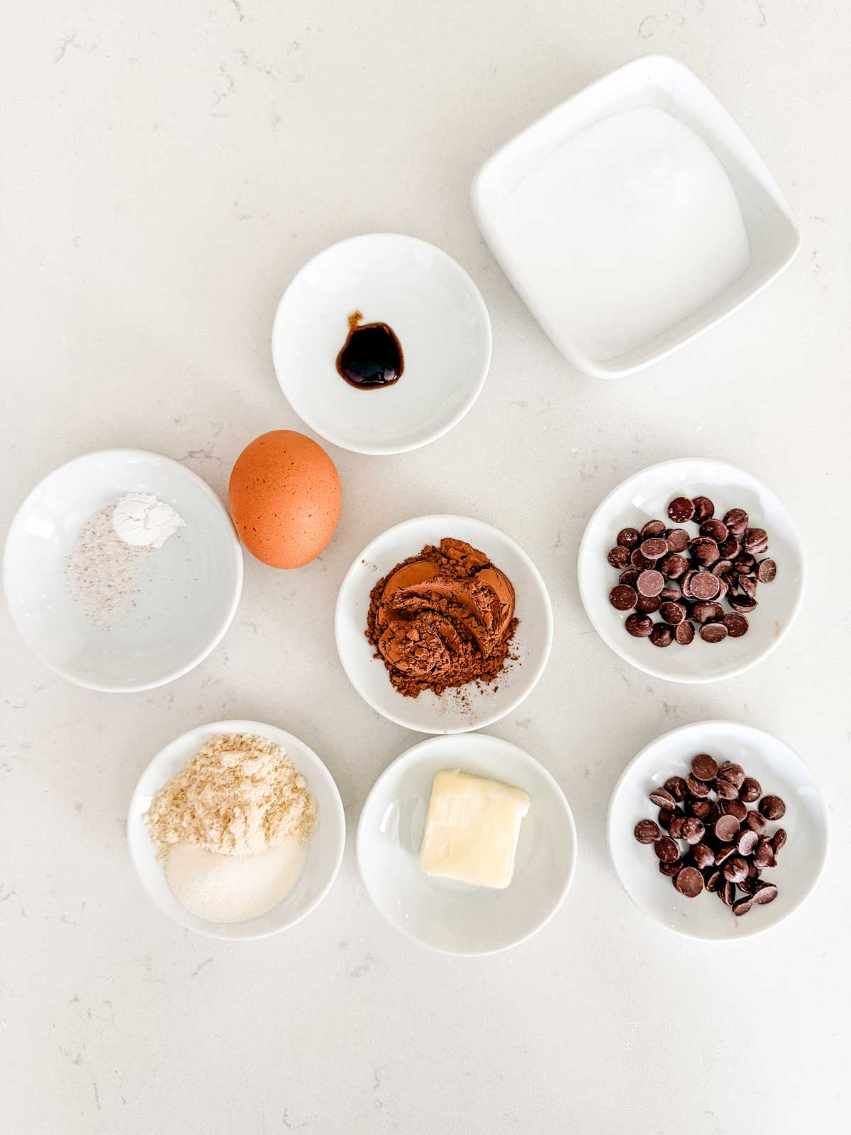 Nine small white bowls and an egg are arranged on a white surface.