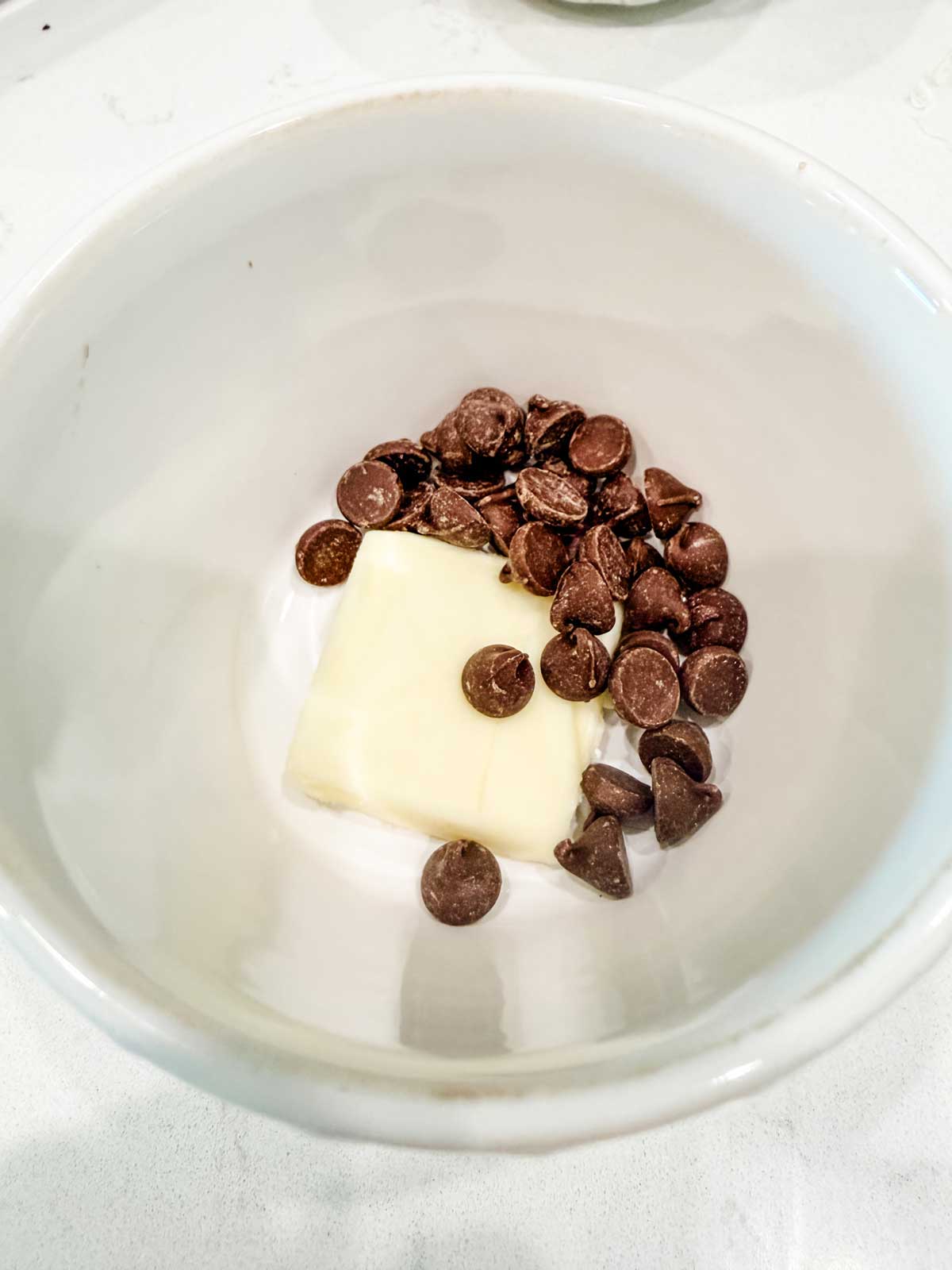 A white bowl containing a small pile of chocolate chips and a rectangular piece of butter on the bottom.