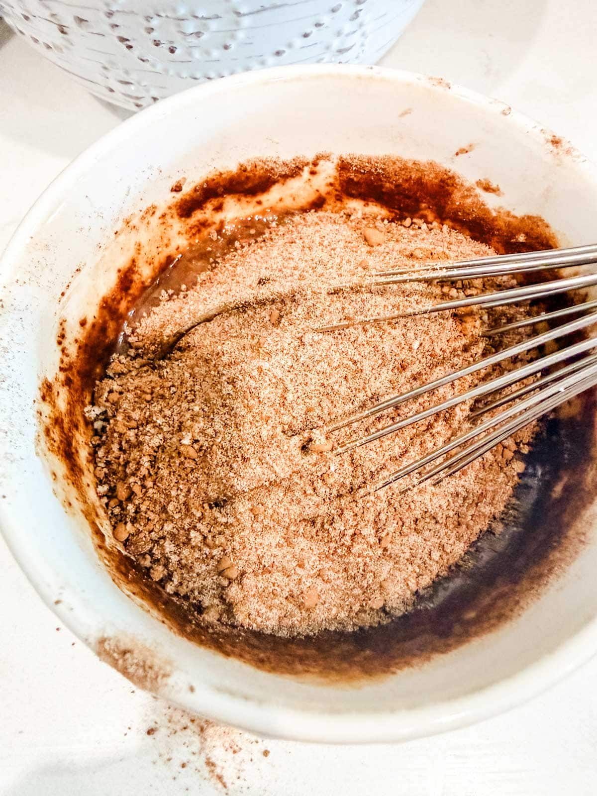 A metal whisk mixes a bowl of dry ingredients, including cocoa powder and flour, in a white bowl on a light surface.