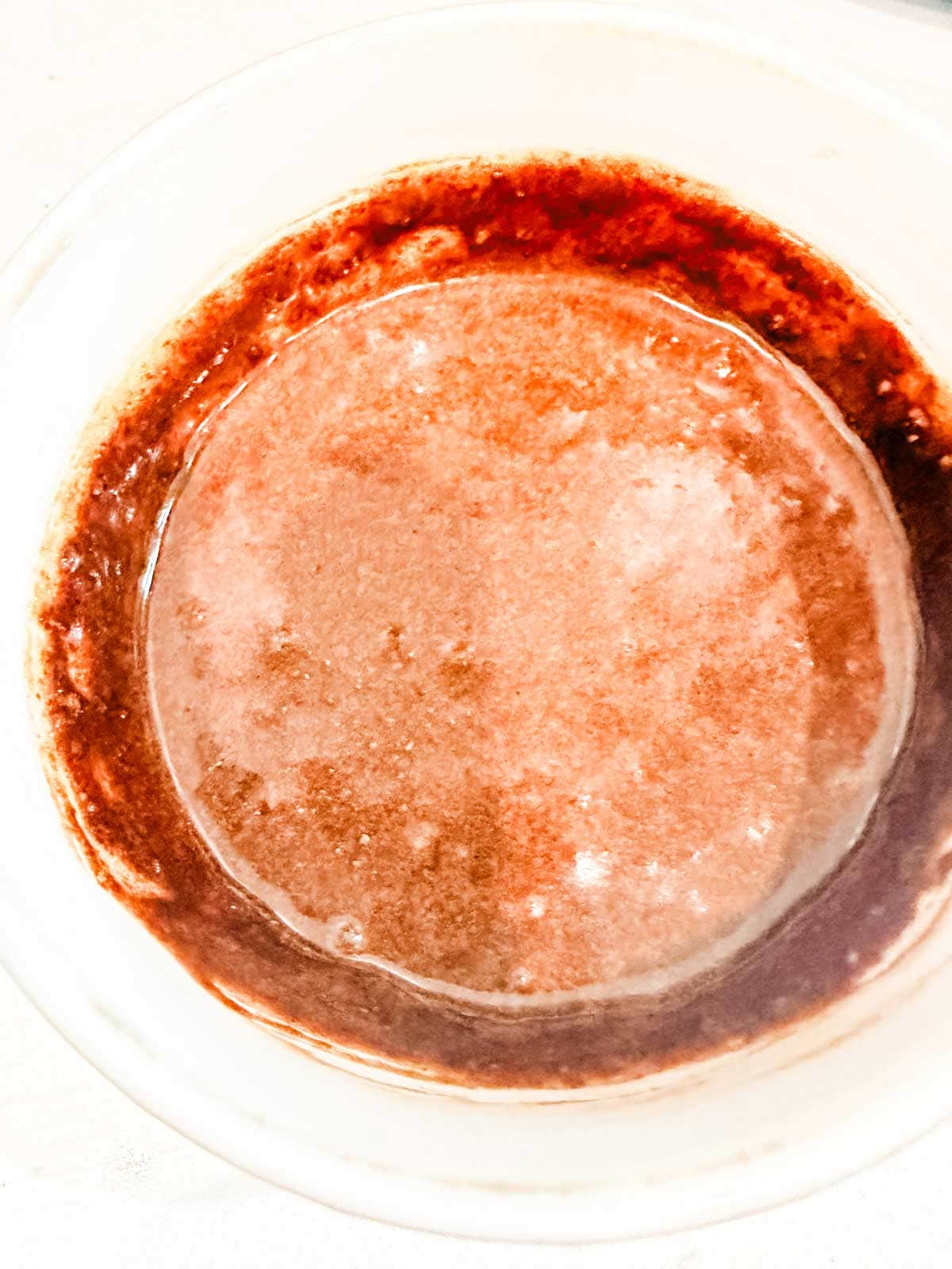 A white bowl filled with a reddish-brown liquid, possibly melted chocolate or sauce, with lighter swirls and patches on the surface.