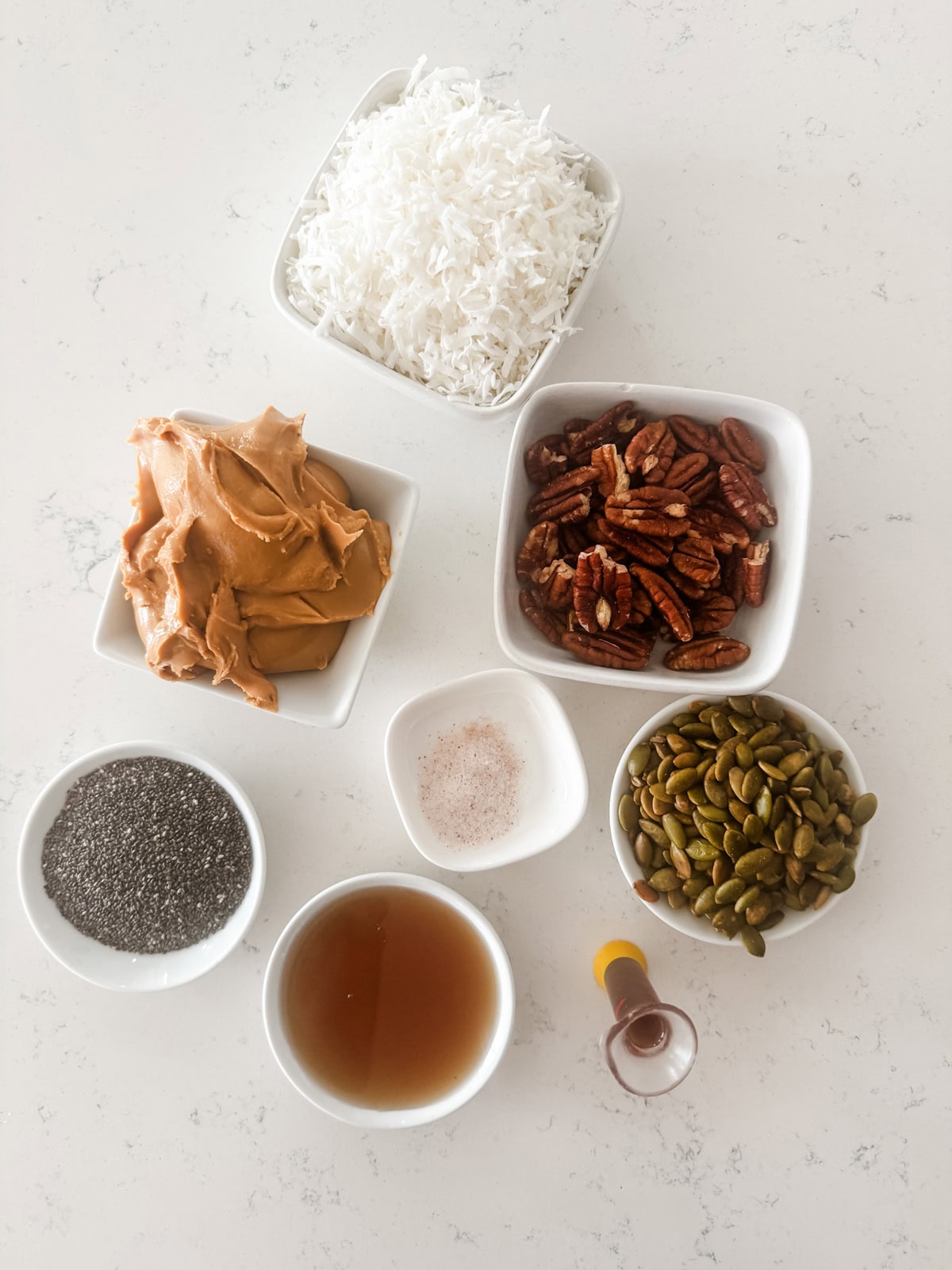 Seven small bowls with ingredients on a white surface: shredded coconut, pecan halves, pumpkin seeds, chia seeds, almond butter, maple syrup, a small vial of vanilla, and a pinch of salt.