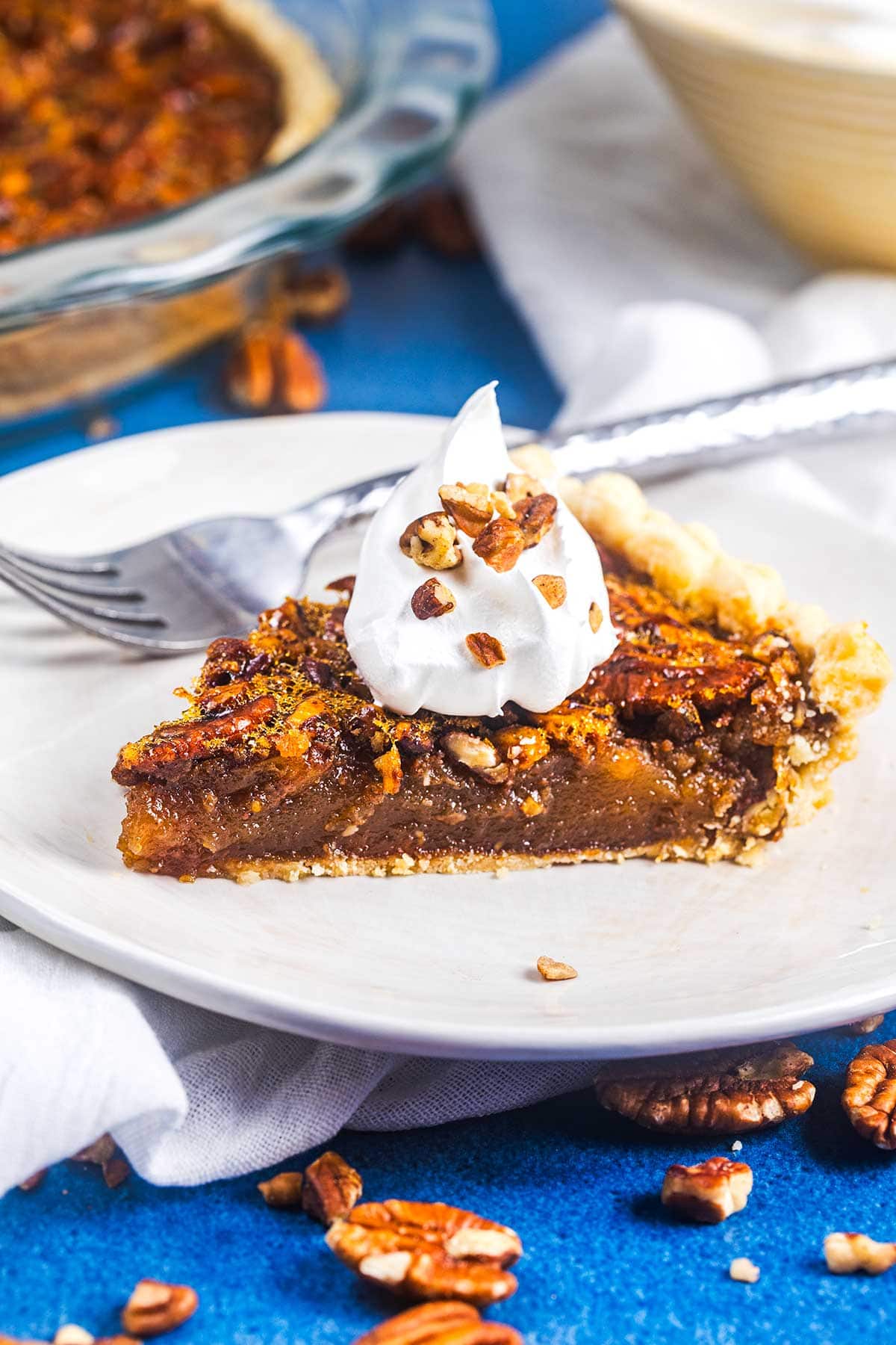 A slice of pecan pie topped with whipped cream and chopped pecans sits on a white plate, with a fork and scattered pecans nearby. The background includes part of the pie in a glass dish and a white napkin.
