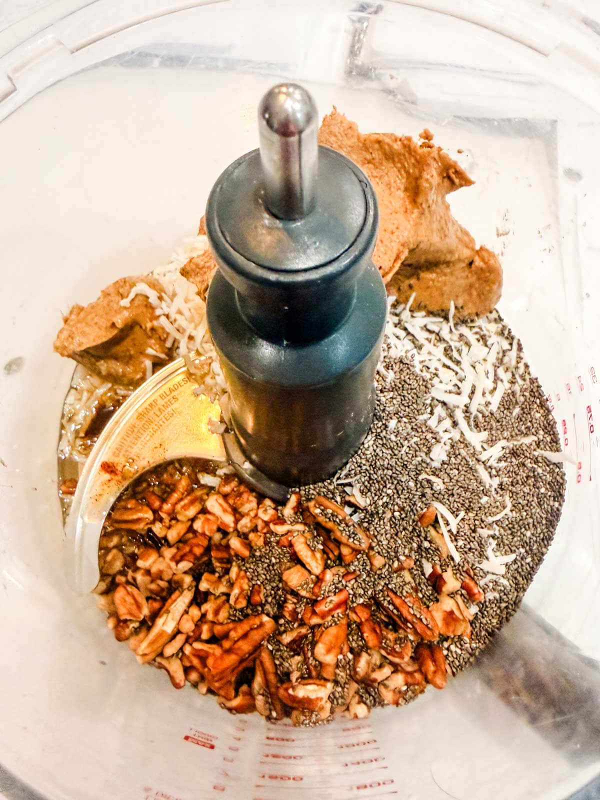 A food processor bowl filled with almond butter, shredded coconut, chia seeds, chopped pecans, and a sticky syrup, ready to be blended together.