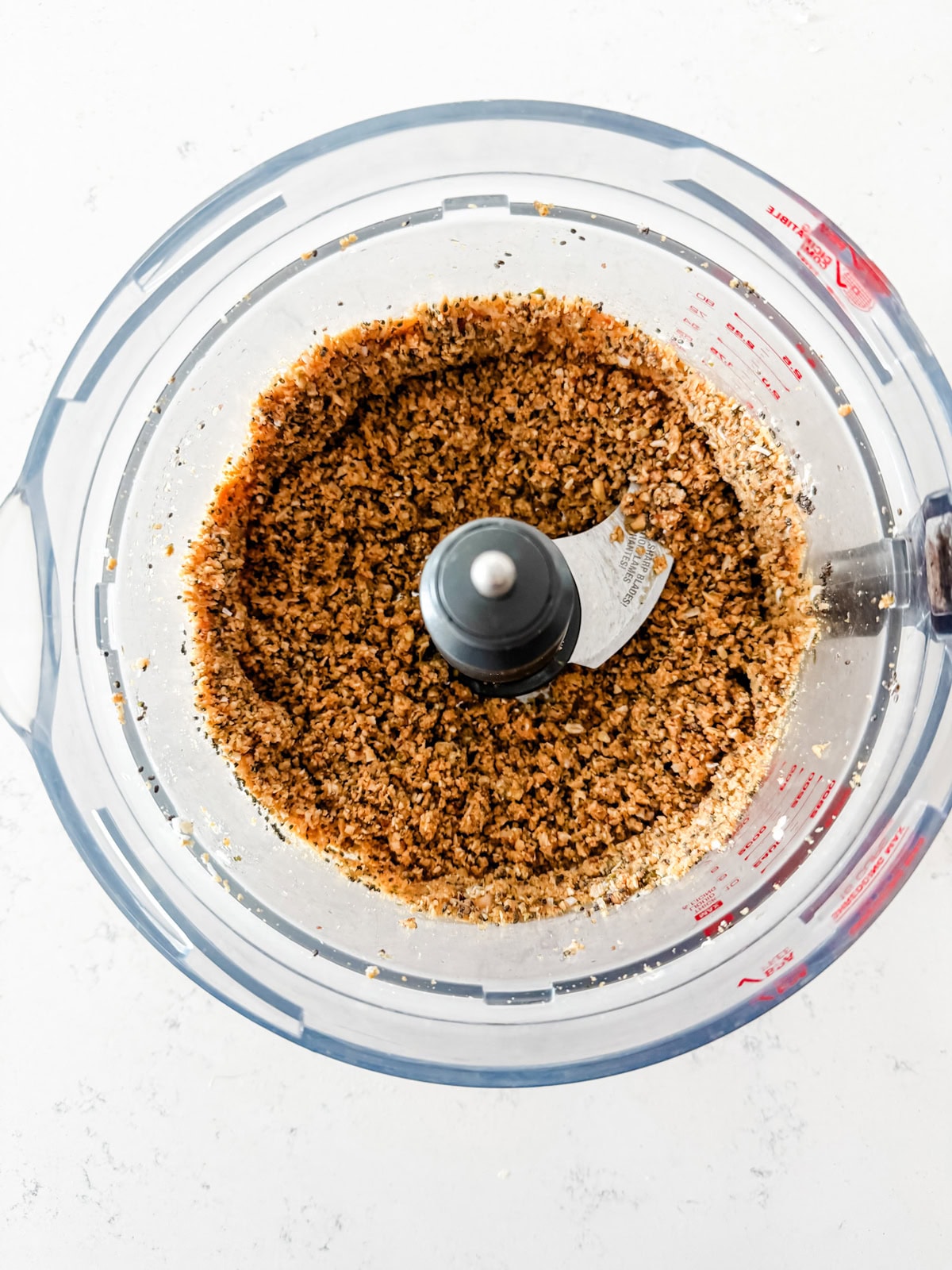 A food processor filled with finely ground, brown and tan mixed ingredients sits on a white countertop, viewed from above.