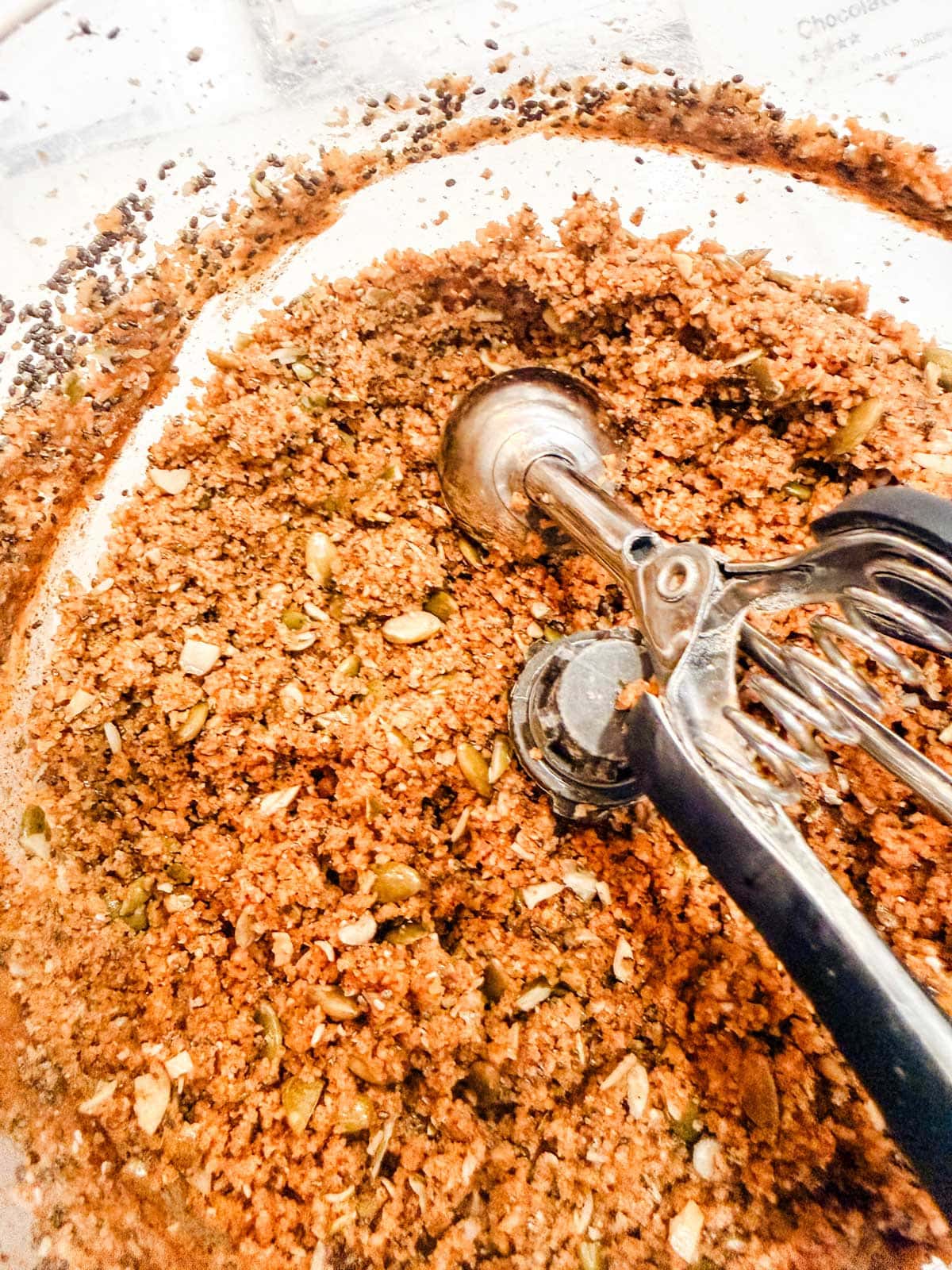 A close-up of a mixing bowl filled with a coarse, brown cookie or granola mixture, with chopped nuts and seeds visible. A metal ice cream scoop rests on top of the mixture.