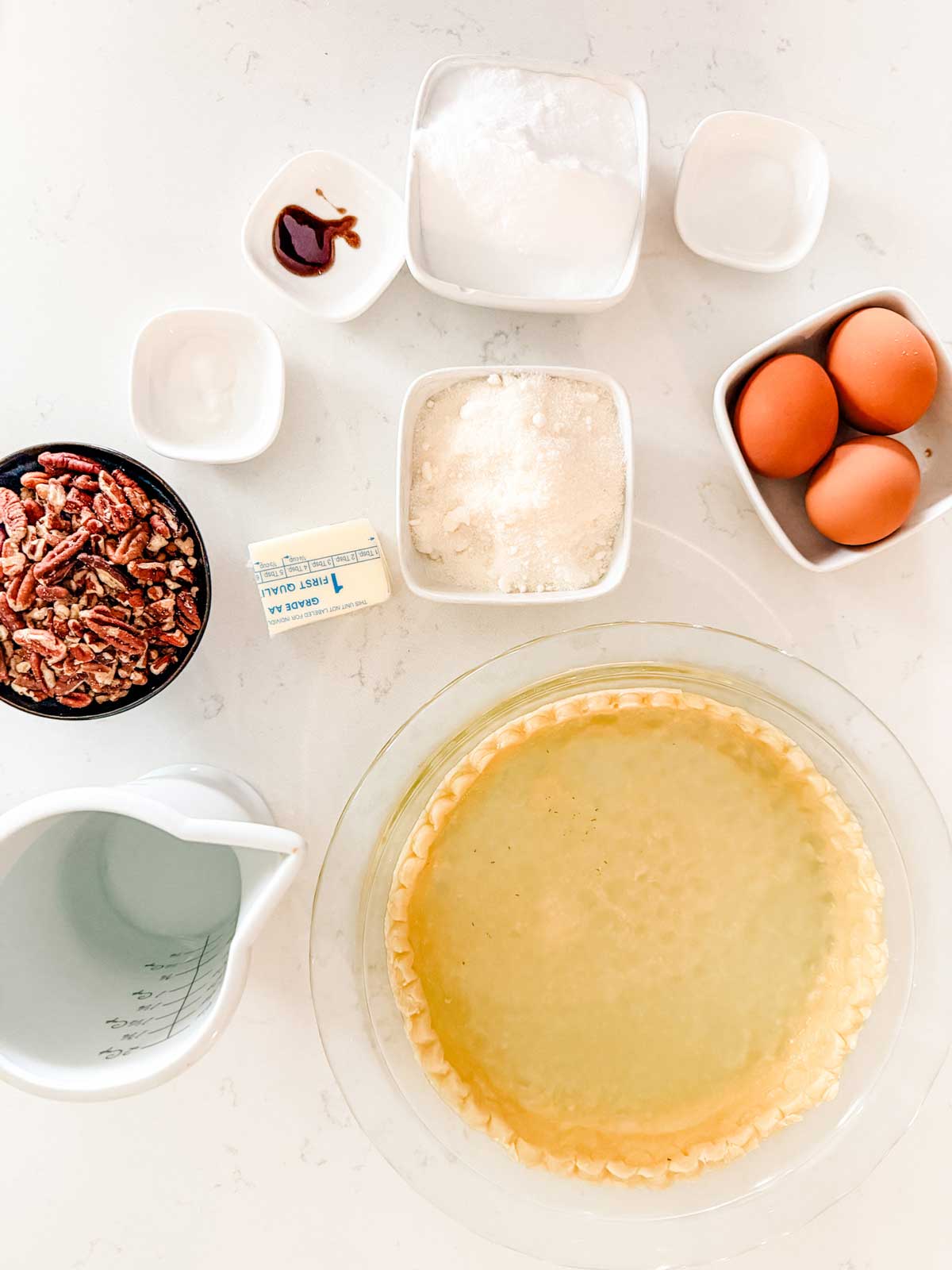 Top-down view of pecan pie ingredients, including a pie crust in a glass dish, pecans, butter, eggs, sugar subsitute, vanilla extract, and a measuring cup of liquid, all arranged on a white surface.