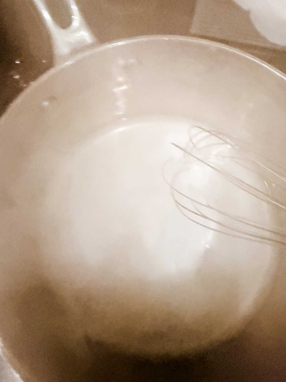 A metal whisk rests inside a large pot filled with a light-colored liquid seen from above; the scene appears hazy, due to steam.