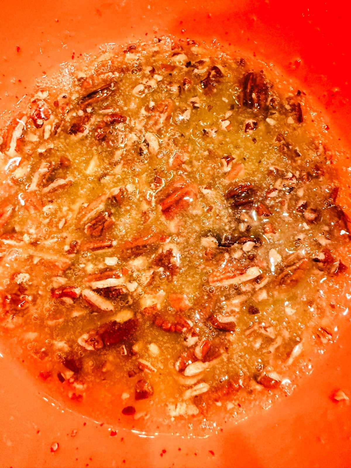 Close-up of chopped pecans mixed in a yellowish, melted butter and sugar mixture inside a mixing bowl.