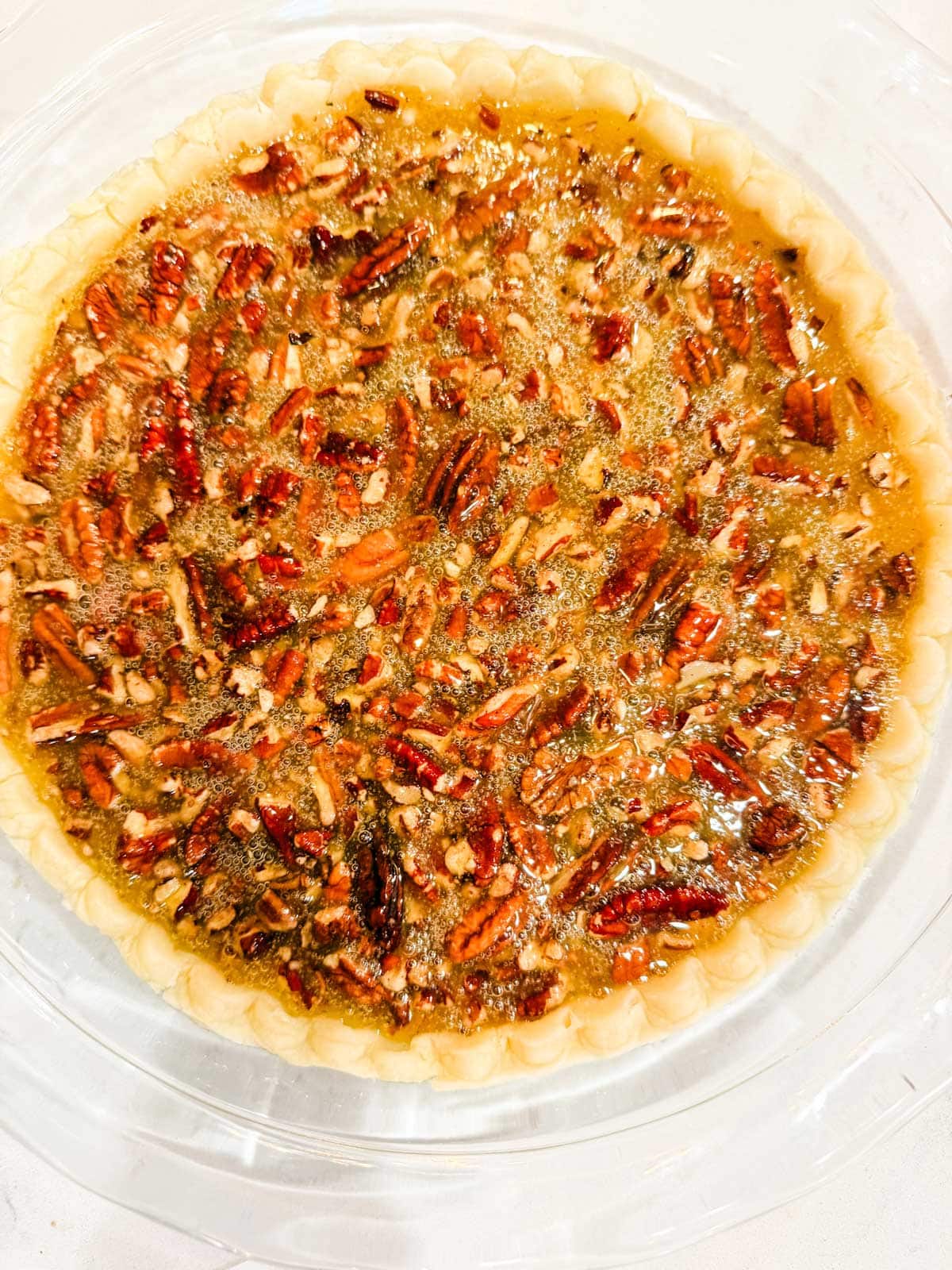 A close-up of a pecan pie with a golden crust and a filling topped with chopped pecans, displayed in a clear pie dish.
