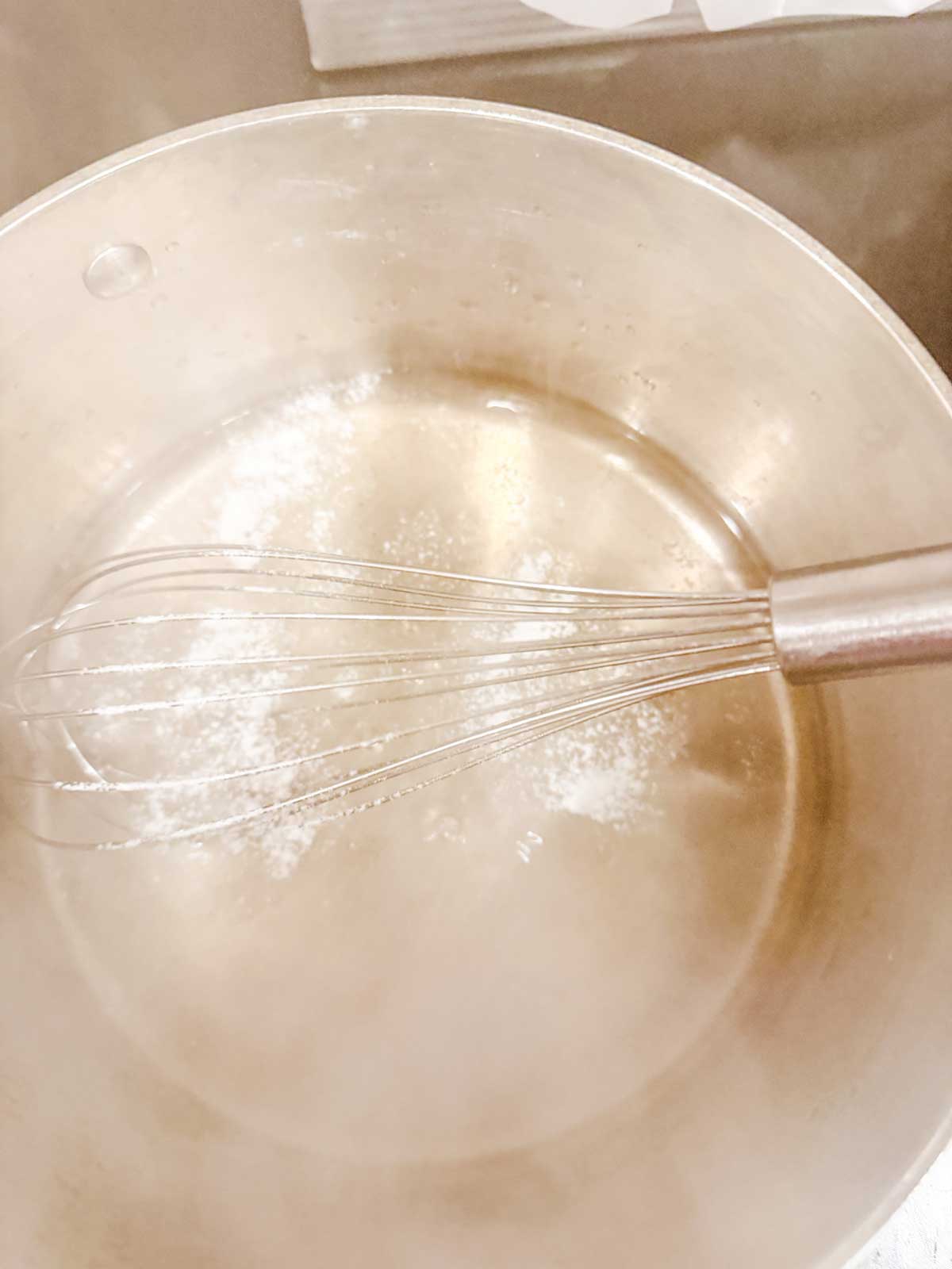 A metal whisk stirring a small amount of xanthan gum in a stainless steel saucepan with water.