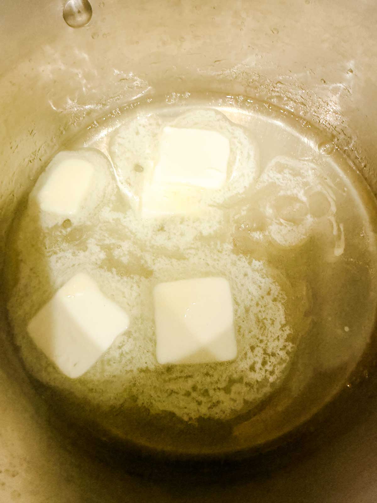 Three pats of butter melting in a saucepan, with some butter already melted and bubbling around the edges.
