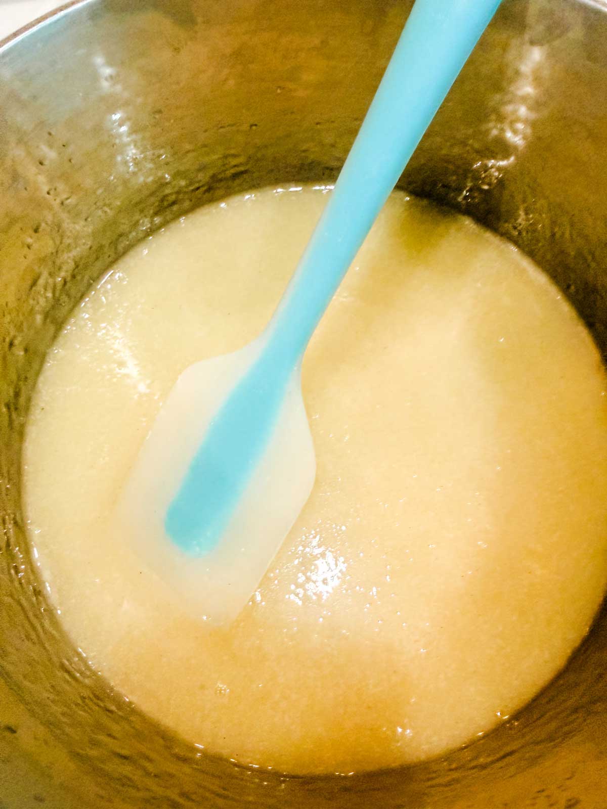 A blue and white silicone spatula stirs a thick, pale yellow mixture in a metal saucepan.