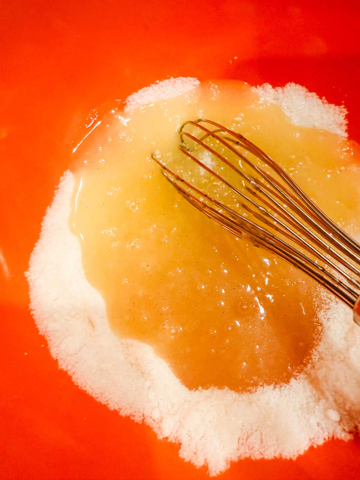 A metal whisk mixing wet and dry ingredients in an orange bowl.