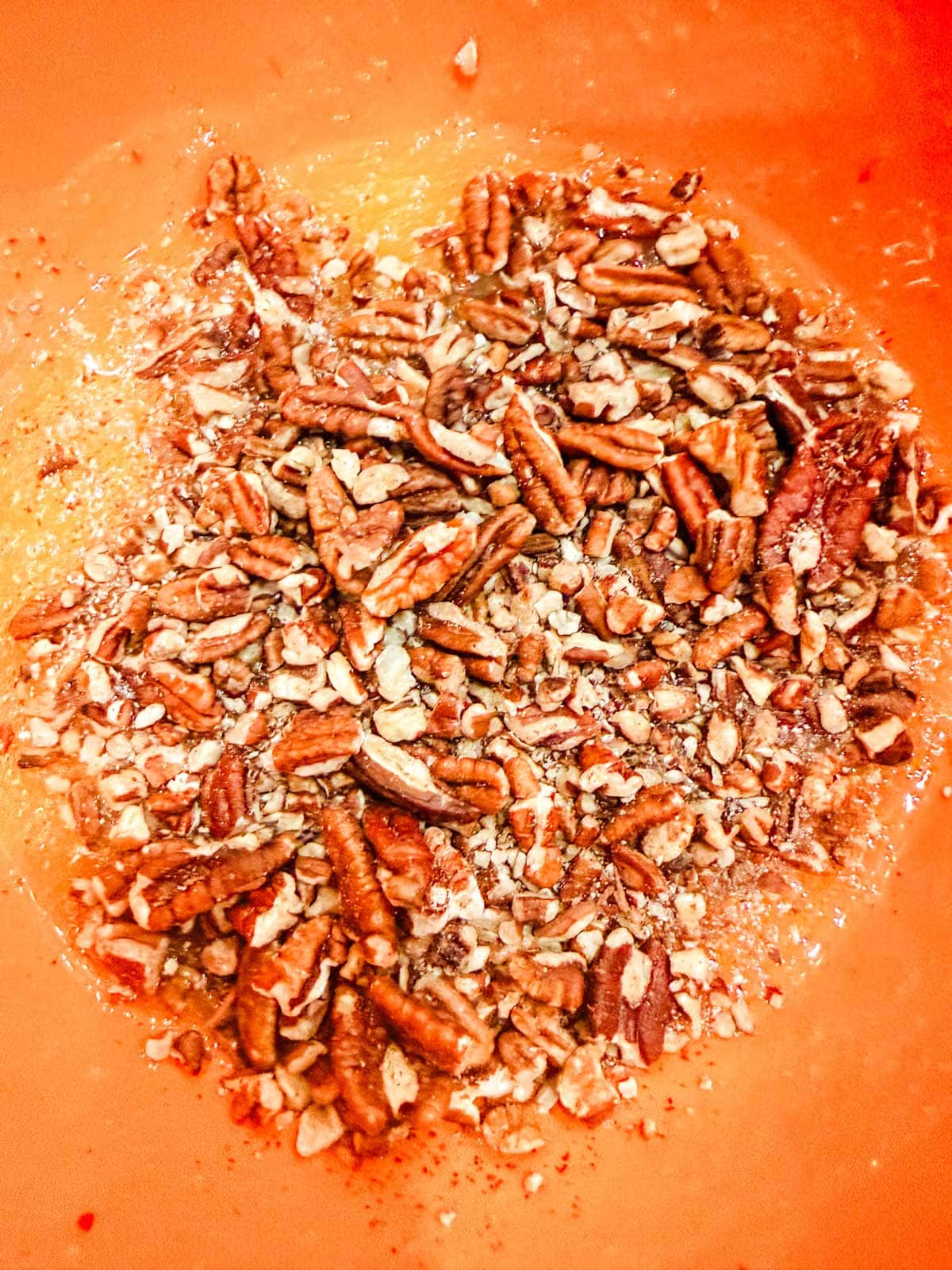 Chopped pecans mixed with a liquid mixture in an orange mixing bowl, creating a textured, sticky mixture.