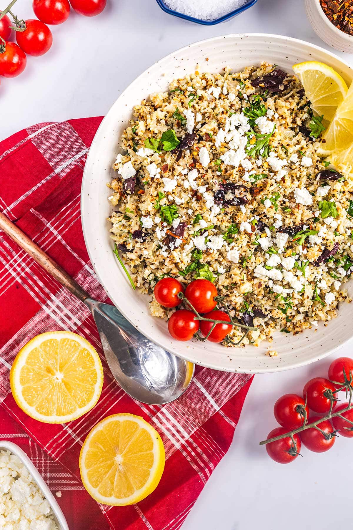 A bowl of cauliflower couscous salad with feta, olives, herbs, and lemon wedges sits on a red checkered cloth, surrounded by cherry tomatoes, lemon halves, and a spoon.