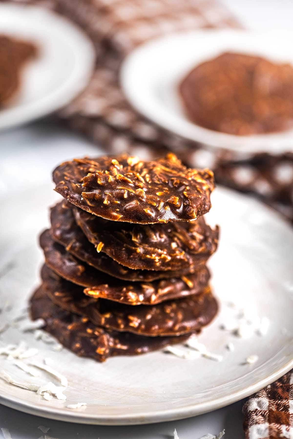A stack of chocolate coconut no-bake cookies sits on a white plate with shredded coconut scattered around, and more cookies are visible on plates in the background.