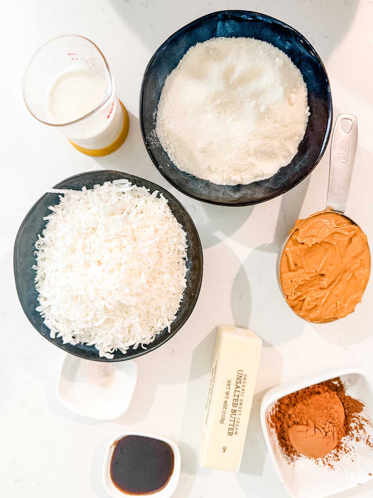 Ingredients for baking are arranged on a white surface, including bowls of keto sweeteners, shredded coconut, peanut butter, a stick of butter, cocoa powder, vanilla extract, cream, and a small dish of salt.