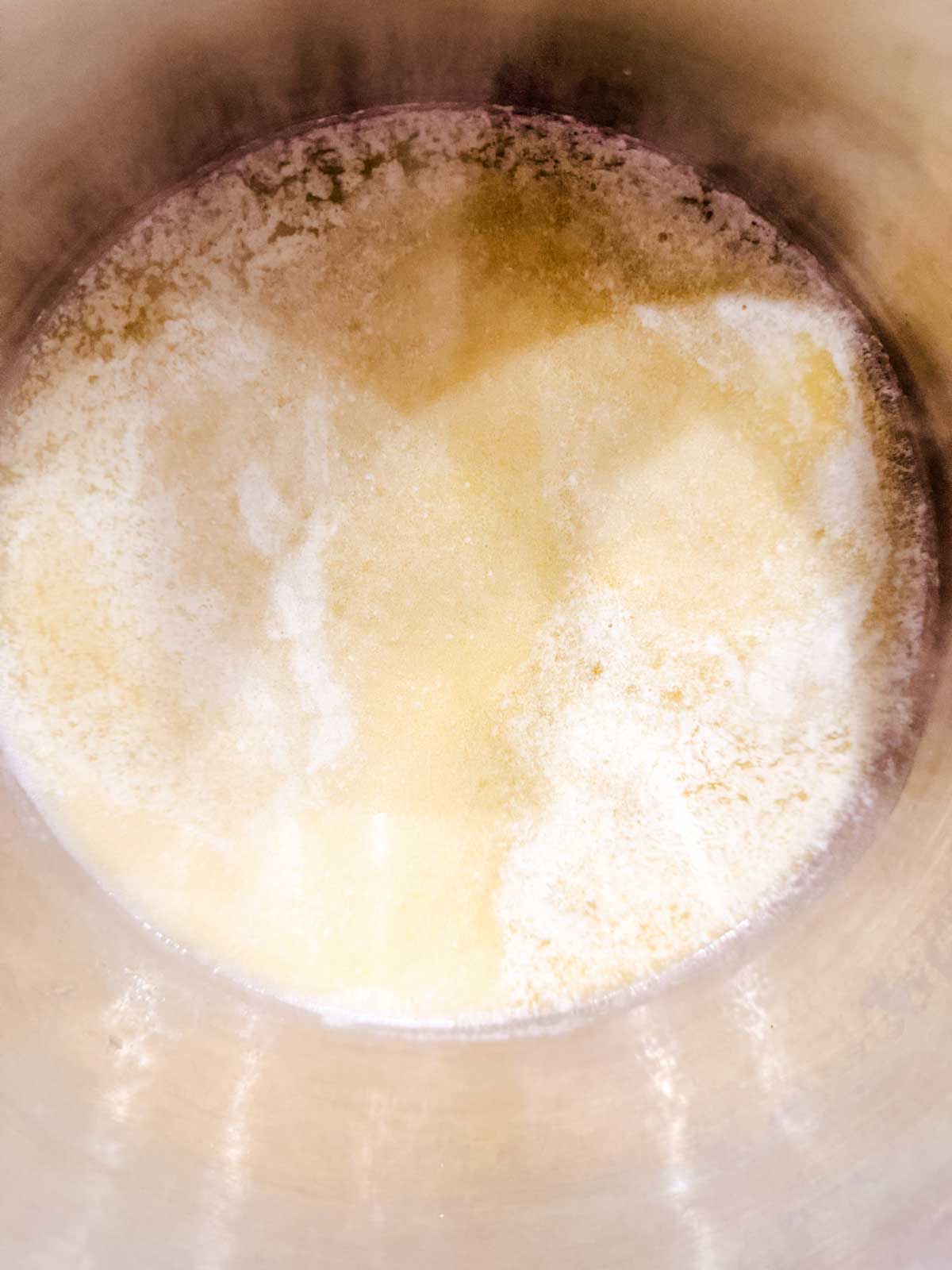 A close-up view of melted butter with milk solids separating in a metallic pan, showing a yellowish liquid with foamy white patches.