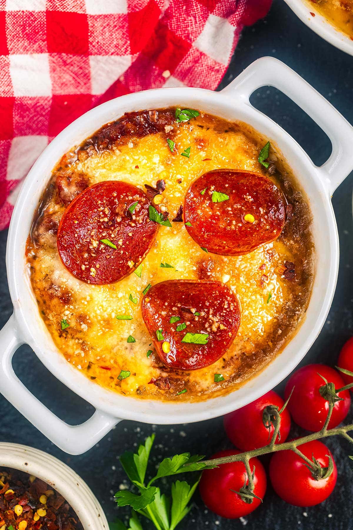 A white ramekin filled with melted cheesy dip, topped with three slices of pepperoni and sprinkled with herbs, sits on a dark surface next to fresh cherry tomatoes and a red-checkered cloth.