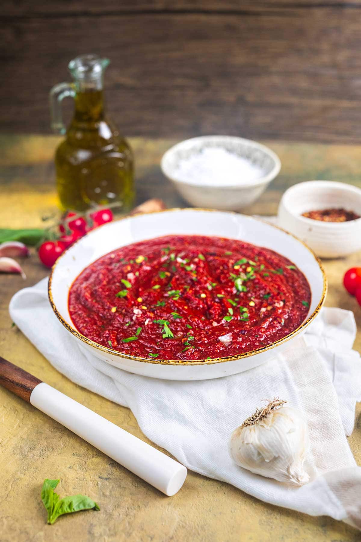 A bowl of red beet dip garnished with herbs sits on a white cloth, surrounded by garlic, radishes, a bottle of olive oil, a bowl of salt, and a small bowl of chili flakes.