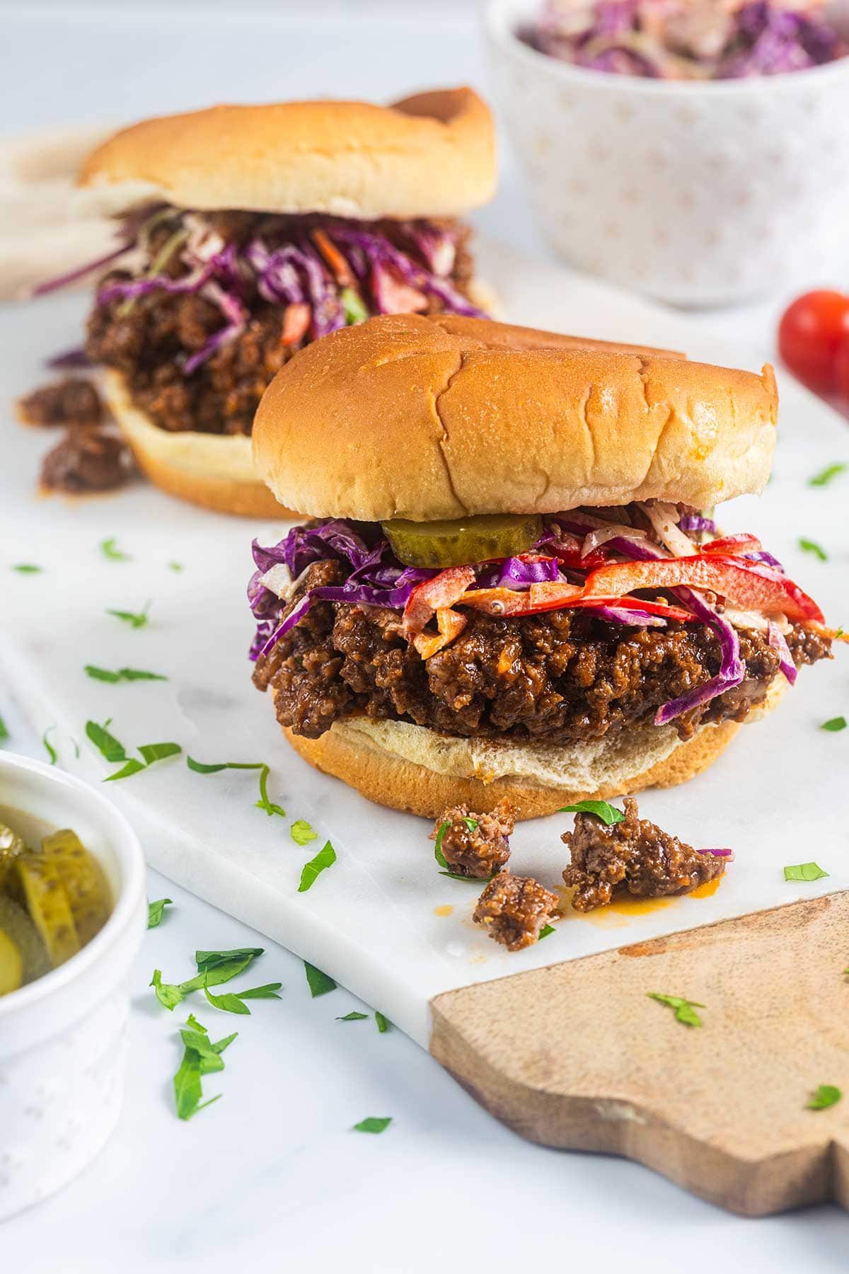 Two sloppy joe sandwiches topped with colorful coleslaw are placed on a white surface. Some filling has spilled onto the surface, and sliced pickles and parsley are nearby. A bowl with more coleslaw is in the background.