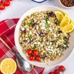 A bowl of cauliflower couscous salad topped with crumbled feta, fresh herbs, olives, and lemon wedges, surrounded by cherry tomatoes, a halved lemon, a spoon, and a red plaid napkin.